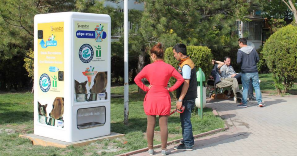image of a recycling bottle machine that gives out dog food in a park with some humans around it